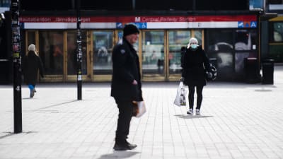 En man i förgrunden och en kvinna med munskydd i bakgrunden som går bort från en metrostation i Helsingfors.
