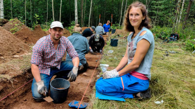 En man och en kvinna sitter vid ett sanddike vid en arkeologisk utgrävning.