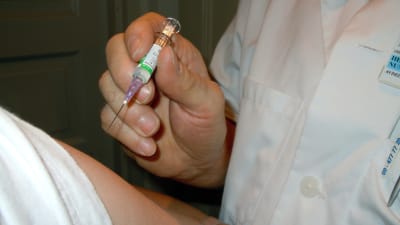 En läkare i vit rock håller i en arm, i den ena handen har hen en vaccinationsspruta.