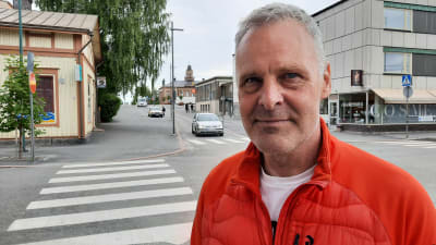 En man i orange jacka tittar rakt in i kameran, där han står på en trottoar i Jakobstads centrum. 