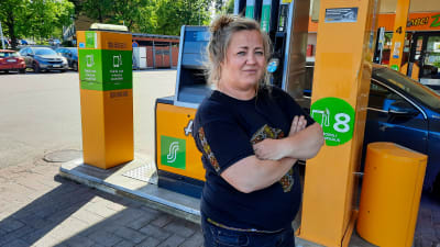 En kvinna står med händerna i kors framför en tankningspunkt på en bensinstation,