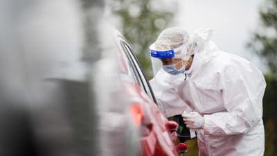 En person utför ett coronatest på en patient som sitter i sin bil (endast bilen syns på bilden). Persoenn som utför testet har skyddsdräkt på sig.