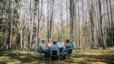 Kuvan etualalla kuusi henkilöä istuu pöydän ääressä ulkosalla kahvilla. Taustalla keväinen koivikko ilman lehtiä.