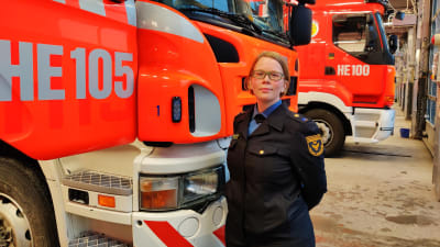 En kvinna med kort blont hår står framför en brandbil i ett stall med flera brandbilar i bakgrunden.