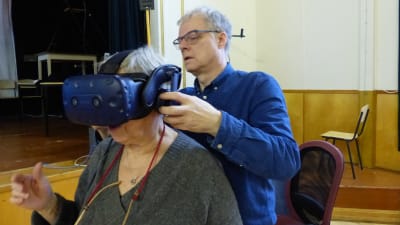 En kvinna sitter i en stol medan en man klär på henne ett par VR-glasögon.