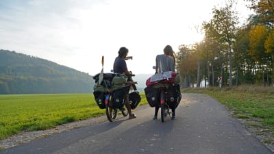 Två cyklister har stannat på en väg och blickar framåt. Vi ser dem bakifrån. Den ena pekar på något. Till vänster syns ett berg. Cyklarna är fullastade med väskor. 