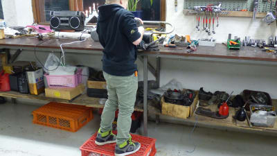 en ung pojke står på en plastpall för att kunna nå upp till verkstadsbordet. Står med ryggen till och blästrar en mopeddel