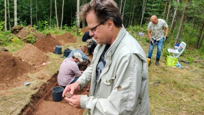 En man står vid en arkeologisk utgrävning och håller i två lerbitar.