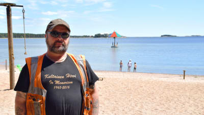 En bild på en man i reflexväst, keps och solbrillor. Han är klädd för arbete och står på en strand som han fixat upp. I bakgrunden syns hav och en vattenkarusell.
