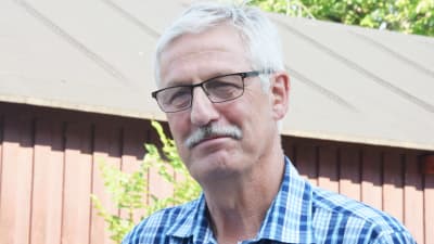 En man med glasögon och blårandig skjorta. Han har mustasch och vitt hår.