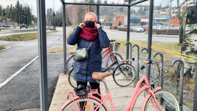 En kvinna sätter på sig ett munskydd. Hon står utomhus vid en cykelställning.