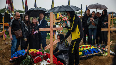 En samling människor med paraplyer på en begravningsplats omgivna av träkors.