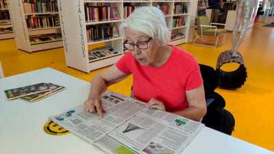 En dam i röd skjorta sitter vid ett bord i ett bibliotek och läser en tidning.