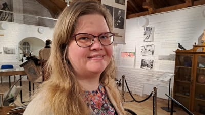 En kvinna står bland olika museiföremål. Hon heter Laura Lotta andersson.