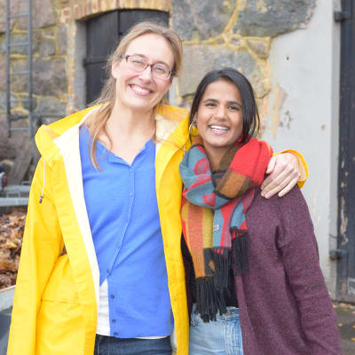 Två kvinnor, en i gul regnrock andra med rutig halsduk. Den med gul rock är längre och äldre och håller sin ena arm om den andra. De är glada och ler stort.