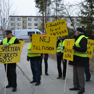 Nio personer står med gula plakat med sloganer mot en kommunsammanslagning.