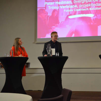 En man och två kvinnor står vid varsitt svart podie på en scen med en 