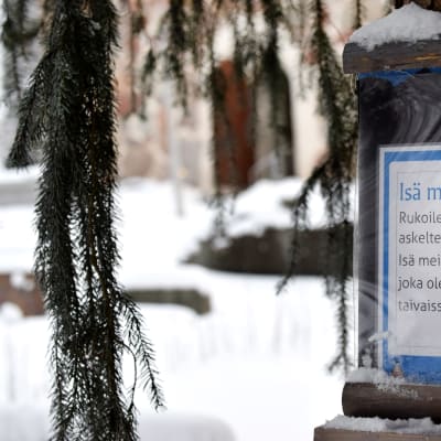 Puunrunkoon kiinnitetty laminoitu kyltti jossa kehotetaan rukoilemaan Isä meidän samalla kun kävelee.