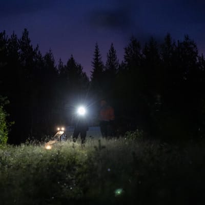 Metsästäjät pimeässä maastossa otsalampun kanssa. Taustalla näkyy pakettiauto.