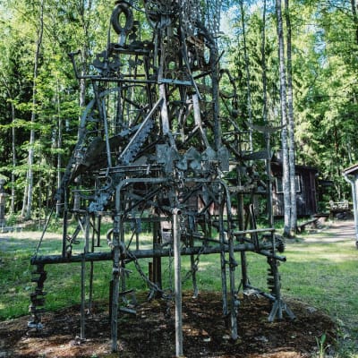 Konstverk gjord av träpinnar, metallrör, sågblad och kedjor ute på en gårdsplan med skog i bakgrunden.