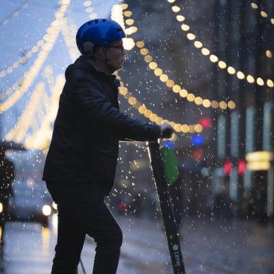 En elsparkcykelåkare med blå hjälm står i skymningen i regn vid Alexandersgatans julbelysning i Helsingfors.