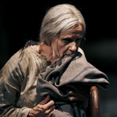 En gammal kvinna i ett smutsigt linne kramar en grå filt.  