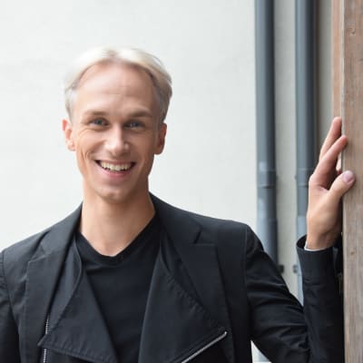 en man i svart läderjacka. ler mot kameran. Lutar sig lätt mot en träpelare. Vit bakgrund.