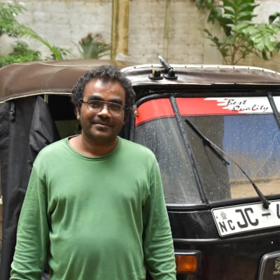 En lankesisk man i grön tröja står framför en svart Tuktuk.