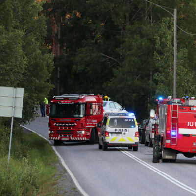 Två stora röda brandbilar och en huvudsakligen vit polisbil på en smal väg omgiven av träd och en åker.  Brandkåren och polisen på olycksplatsen i Borgå.