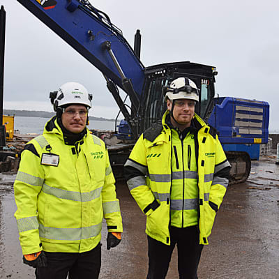 Två byggnadsarbetare står framför en grävmaskin.