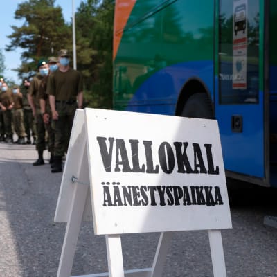 en vit skylt där det står "vallokal", i bakgrunden står beväringar och köar för att få rösta. 