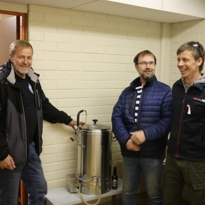 Tre män står vid en ölbryggartunna.