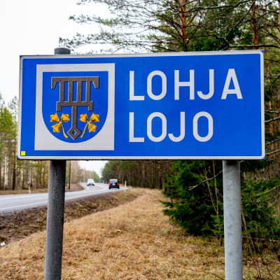 En blå skylt i terrängen vid kommungränsen där det står Lojo/Lohja.