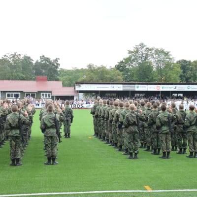 Militärparad med rekryter ute på en gräsplan. I bakgrunden pubilk.