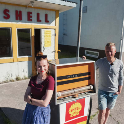 Man och kvinna lutar sig mot gammal bensinpump. I bakgrunden en liten kiosk med gula knutar samt texten SHELL.