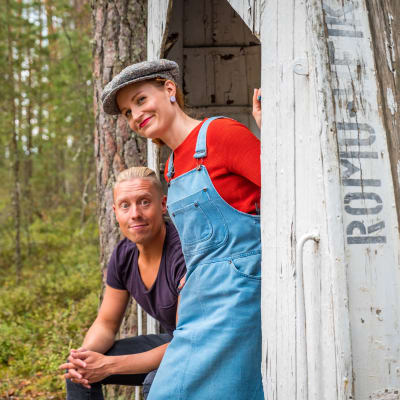 Egenlands programledare Hannamari Hoikkala och Nicke Aldén på ön Hytermä i Kerimäki - brevid en bänk som är gjord av en båt som lyfts upp. På båten står det: Romu-Heikki.