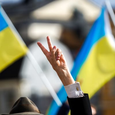 Henkilö näyttää voitonmerkkiä sormillaan Ukrainan lippuja taustalla.