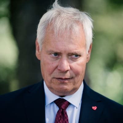 Antti Rinne i mörk kostym och röd slips mot en grön suddig bakgrund. Han har en sammanbiten min.