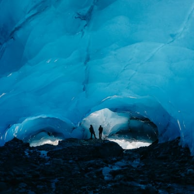 Fredrik och Katja står inne i grottan. Det är stenar på marken och glaciärtaket är som ett stort valv i turkost. 
