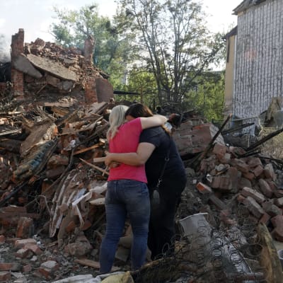 Kaksi naista halaa taustallaan tuhoutuneita asuinrakennuksia.