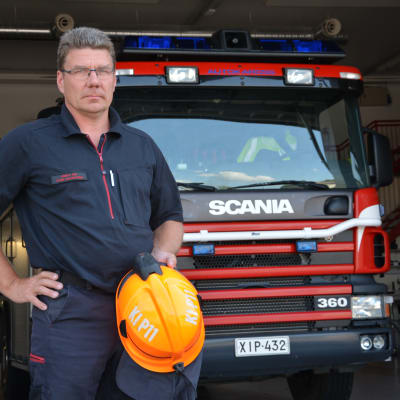 Sture Holmström med en orange hjälm i handen framför en brandbil.