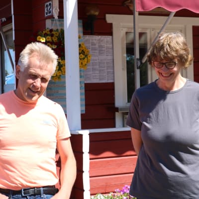 Ett medelålders par står vid en röd stuga. Mannen har en orange t-skjorta, kvinnan en blågrå.