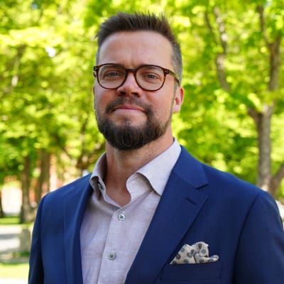 En man med mörkt hår, glasögon och mörkblå kostym ler mot kameran.