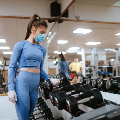 En kvinna i munskydd lyfter upp vikter på ett gym.