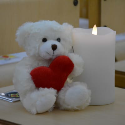 En vit nallebjörn med ett hjärta i famnen sitter intill ett ljus. 