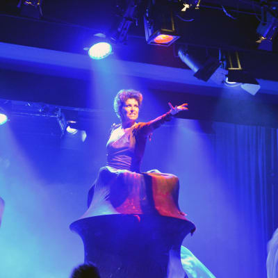 En kvinna på scen som står inne i ett "berg" med den enda armen utsträckt mot publiken.