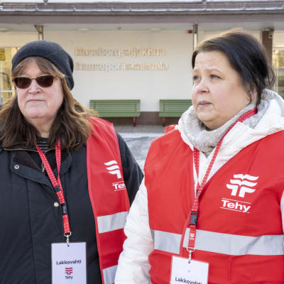 Två kvinnor i vinterkläder och rödvita västar utanför ett sjukhus.