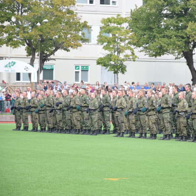 Militärparad med rekryter ute på en gräsplan. I bakgrunden vita byggnader och träd.