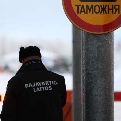 Henkilö, jonka takissa teksti Rajavartiolaitos ja päässä karvalakki seisoo selin kameraan, vieressään liikennemerkki, jossa kirjoitusta kyrillisin kirjaimin.