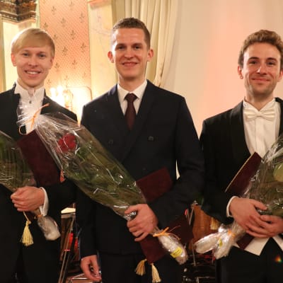 Kolme miestä seisoo kukkakimput käsissään juhlapuvuissa.